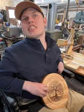 Karl at his business holding one of his designs, a circular piece of wood with an intricate carving of a tree on it