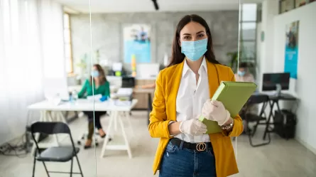 Female office worker wearing yellow jacket and face mask standing outside an office with a person sitting at a desk wearing a face mask.