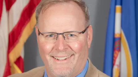 Tim, wearing a dress shirt and suit jacket in front of U.S. and Minnesota flags