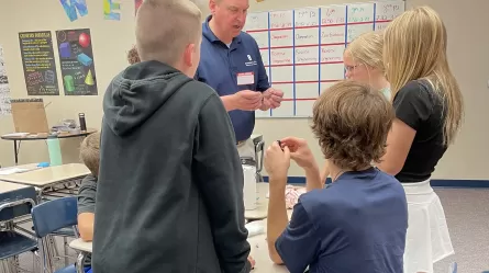 Adult instructor and teenage students standing around a table working on a project in a classroom.