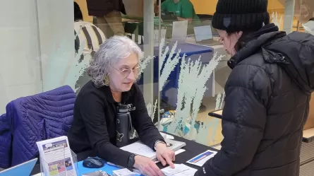 Sandra Brick, a person sitting at a table with brochures and other materials, helping a person wearing a hat and jacket standing at the table