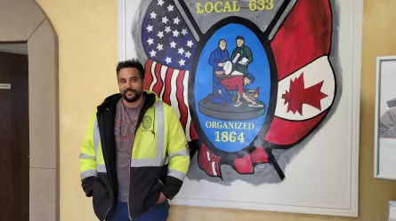 a person in a yellow and black jacket with reflective strips standing in front of a sign with the U.S. and Canadian flags that reads Local 633 organized1864