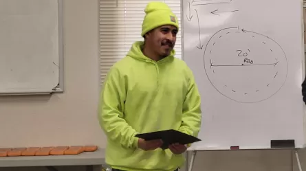 person in bright yellow hoodie and cap holding a certificate standing in front of a white board
