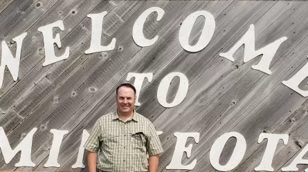 a person wearing a plaid shirt in front of a wooden sign that reads Welcome to Minneota