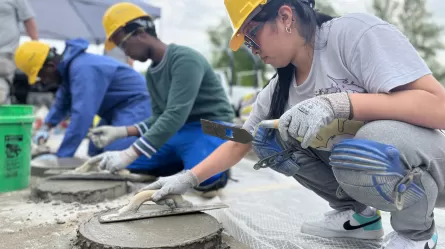 adults participating in cement masonry training, people are wearing helmets and eye protection and using tools to smooth cement surfaces