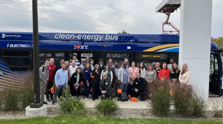 many people standing in front of a bus with writing on it that reads New York City Bus and electric bus and clean energy bus