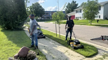 two people standing outside on a lawn, one is pushing a lawnmower, the other has a lawn trimmer in this hands