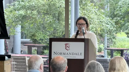 person speaking into a microphone, Normandale Community College logo on the lectern,Mue Hsae speaking at semiconductor manufacturing program graduation June 2025 