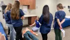 four people standing with their backs to the camera looking at an instructor demonstrating care on a mannequin on the floor