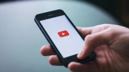 Person's hand with a cell phone. On the cell phone screen their is a red play video icon.