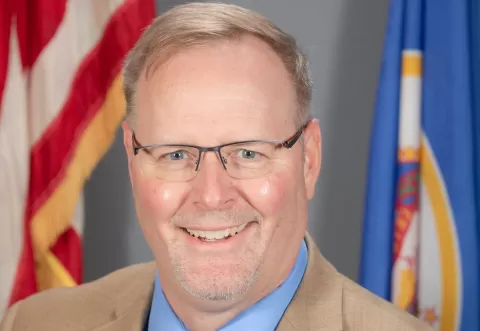 Tim, wearing a dress shirt and suit jacket in front of U.S. and Minnesota flags
