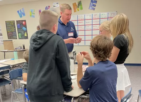 Adult instructor and teenage students standing around a table working on a project in a classroom.