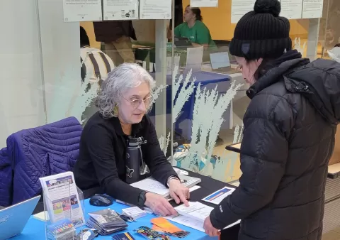 Sandra Brick, a person sitting at a table with brochures and other materials, helping a person wearing a hat and jacket standing at the table