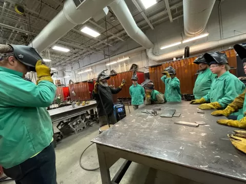 several people wearing welding helmets and protective clothing standing around a metal table in a machine shop