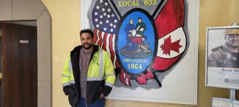 a person in a yellow and black jacket with reflective strips standing in front of a sign with the U.S. and Canadian flags that reads Local 633 organized1864
