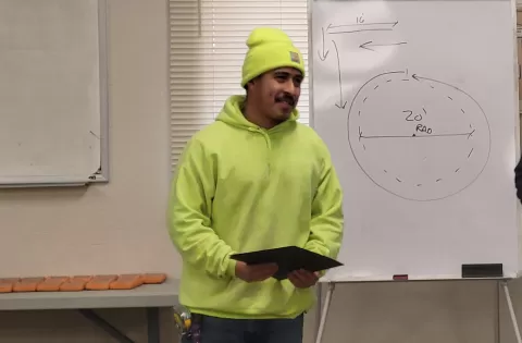 person in bright yellow hoodie and cap holding a certificate standing in front of a white board