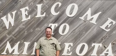 a person wearing a plaid shirt in front of a wooden sign that reads Welcome to Minneota