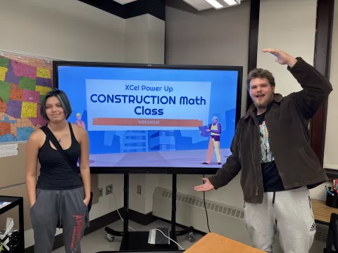 two people standing on either side of a large monitor that reads Xcel Power Up Construction Math Class