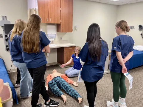 four people standing with their backs to the camera looking at an instructor demonstrating care on a mannequin on the floor