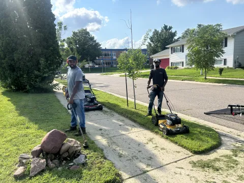 two people standing outside on a lawn, one is pushing a lawnmower, the other has a lawn trimmer in this hands