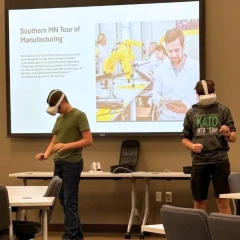 two people using virtual reality goggles in front of a screen with the words 'Southern Minnesota Tour of Manufacturing'