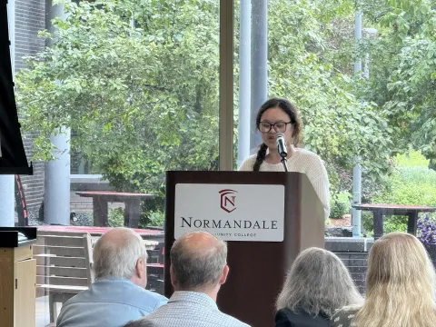 person speaking into a microphone, Normandale Community College logo on the lectern,Mue Hsae speaking at semiconductor manufacturing program graduation June 2025 