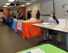people sitting and standing at tables with materials for display arranged around perimeter of a large room