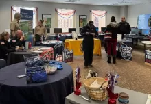 people sitting and standing at tables with materials for display arranged around perimeter of a large room